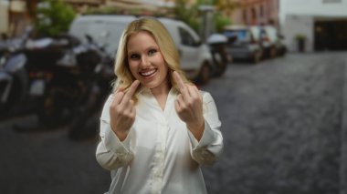 Sarışın genç bir kadın şehir sokaklarında gülümsüyor ve iki parmağı şehir merkezinde park halindeki araçlarla çevrili neşeli hareketler sergiliyor..