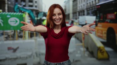 Şehir merkezinde kızıl saçlı, kırmızı kazaklı bir kadın inşaat alanında gülümsüyor canlı enerji ve şehir manzaralı modern gelişimi sergiliyor..