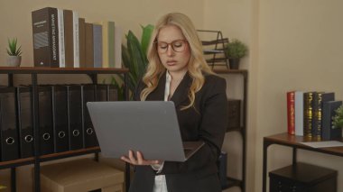 Modern ofis ortamında dizüstü bilgisayarda çalışan bir kadın, kitaplık ve iş kıyafetleriyle profesyonel ve odaklanmış bir atmosfer sergiliyor..