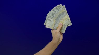 İzole edilmiş bir mavi duvar arkaplanına karşı Endonezya banknotları tutan erkek el, finans ve para değişimi kavramlarını vurguluyor.