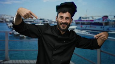 Siyah üniformalı bir şef gibi giyinmiş İspanyol sakallı adam arka planda gemiler ve teknelerle dolu bir limanın yakınlarında neşeli bir şekilde el kol hareketi yapıyor..