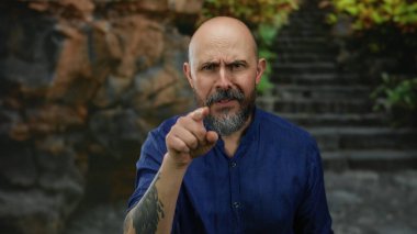 Mavi tişörtlü kel bir adam kayalık bir arka planı olan açık şehir ortamında ciddi bir şekilde el kol hareketi yapar. Yoğun ifadesini ve çekici tavırlarını yakalar..