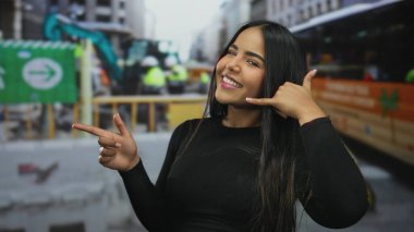 Latin asıllı genç bir mankenin açık havada gülümseyip işaret ettiği bir inşaat alanında el kol hareketi yapan bir kadın..