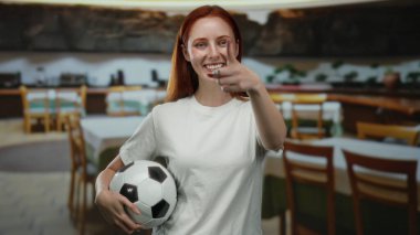 Restoranda futbol topuyla gülümseyen, kamerayı işaret eden, beyaz gömlekli, kırmızı saçlı bir kadın.
