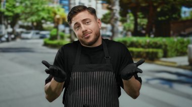 Önlüklü genç sakallı adam eldivenleriyle sokaklarda omuz silkiyor şehir arka planında açık havada garson teması sergiliyor.