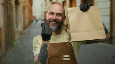 Gülümseyen adam şehir sokaklarında kağıt torba tutuyor. Önlük dövmeleri ve eldivenler takıyor. Bu da modern teslimat servisini işaret ediyor..