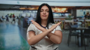 Kapalı bir lokantada kollarını kavuşturmuş beyaz üniformalı bir kadın, hareketli bir kahve dükkanı ortamında kendinden emin görünüyor..