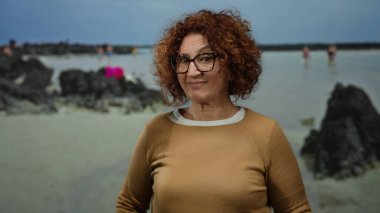 Kumlu sahilde duran, arka planda bulutlu bir gökyüzünün altında kayalar olan, gözlüklü, kıvırcık esmer saçlı, rahat bir deniz havası yakalayan bir kadın.