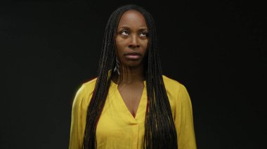 Sarı tişört giyen Afrikalı kadın koyu bir arka plana karşı duruyor, örgülü saç stilini ciddi bir ifadeyle gösteriyor..