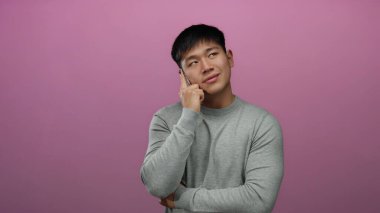 Akıllı telefondan konuşan genç bir adam pembe bir arka plana karşı içerik ifadesiyle gri bir kazak giyiyor ve kapalı bir ortamda sakin bir hava yayıyor..