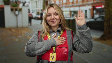 Sarı saçlı genç gönüllü bir kadın şehir sokaklarında kırmızı yelek ve sarı etiket takıyor, açık havada bağlılık gösteriyor..