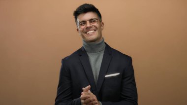 Genç bir adam kendine güvenerek gülerek ve alkışlayarak izole edilmiş kahverengi bir arka plana karşı şık giyiminin ve pozitif davranışının altını çiziyor..