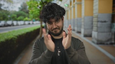Arka planda ağaçlar ve arabalarla şehir sokağında el çırpan sakallı genç İspanyol bir adam sıradan moda ve doğal ifade sergiliyor..