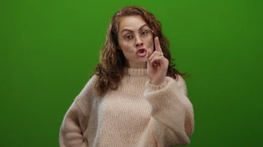 Gözlük takan ve rahat bir süveter giyen bir kadın, izole edilmiş yeşil bir arka plan duvarına karşı anlamlı bir şekilde jest yapar, canlı bir ifade ve kişilik anı yakalar..