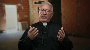 Siyah cüppeli kıdemli rahip, duvarlarla örülü bir inşaat alanında düşünceli bir şekilde el kol hareketi yapar, kırsal bir alanda tefekkür ve kendini adama anını yakalar..