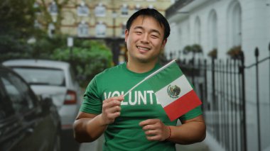 Kentsel caddede Meksika bayrağı taşıyan genç adam gönüllü gömleğiyle gülümsüyor. Kültürel çeşitliliği ve şehir ortamında toplumsal katılımı gösteriyor..