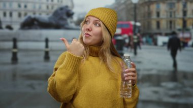 Sarı kazaklı ve bereli bir kadın su şişesi tutuyor. Arka planda bulanık bir parmak izi var. Açık şehir ortamında kendine güveni ve günlük tarzı var..