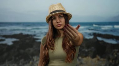 Deniz kıyısında jest yapmaya davet eden kadın, sakin okyanus manzarasına tepeden bakan ciddi bir ifadeyle hasır şapka takıyor.