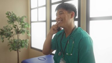 Yeşil önlüklü genç doktor gülümsüyor ve telefonda, parlak bir hastane odasında stetoskop ve kimlik kartıyla konuşuyor..