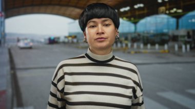 Havaalanında el kol hareketi yapan, çapraz kollar ve sert ifadeyle hoşnutsuzluğunu ifade eden, modern açık hava terminalinde çizgili kazak giyen bir kadın..