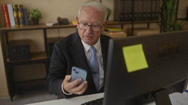 Ofiste bilgisayar başında çalışan kıdemli bir adam, akıllı telefonunu kontrol ediyor, etrafı kitaplar, bitkiler ve kırtasiye malzemeleriyle çevrili, profesyonel bir iş yeri düzeneği kuruyor..