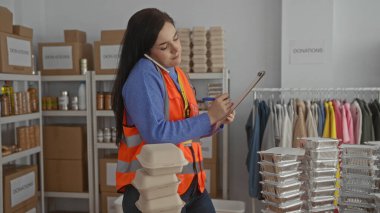 Gönüllü olarak birden fazla iş yapan bir kadın içeride duruyor, telefonla konuşuyor ve arka planda bağış kutuları ve giysi raflarıyla notlar alıyor..