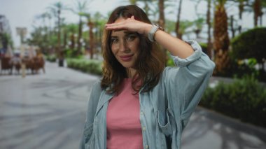 Genç bir kadın güneşli bir parkta dikiliyor. Elleriyle gözlerini koruyor. Palmiye ağaçlarıyla çevrili..