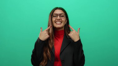 Gülümseyen İspanyol kadın iki eliyle kendini işaret ediyor, gözlük takıyor ve kırmızı gömleğin üzerine siyah bir ceket giyiyor, yeşil arka plan duvarına karşı duruyor..