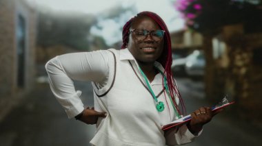 Elinde dosya olan doktor kadın şehir caddesindeki açık havada duruyor. Kızıl saçlı, şehir ortamında düşünceli görünüyor..
