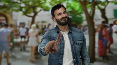 Sakallı İspanyol bir adam, arka planda bulanık insanlar ve ağaçlarla dolu kalabalık bir sokakta dikilirken 