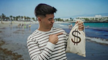Genç adam, arka planda okyanus olan bir kumsalda para çantasını işaret ederek zenginliği, rahatlığı ve kıyı havasını vurgulayan neşeli bir anı yakalıyor..