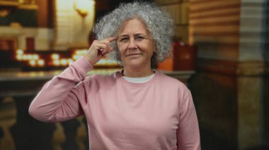 Kilise ortamında el kol hareketi yapan, düşünceli bir ifadeyle tuhaf bir jest yapan, gri bukleli ve pembe giyimli bir kadın ev ortamında ilgi çekici bir kontrastlık yaratır..