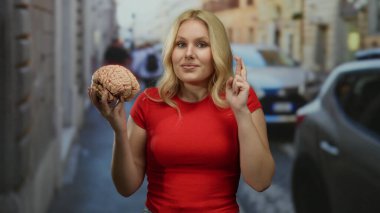 Şehirdeki sokak ortamında parmaklarını çaprazlarken beyni tutan kadın kırmızı tişört giyip şehir çevresindeki düşünce ve umudu sembolize ediyor. Etrafta arabalar ve binalar var..