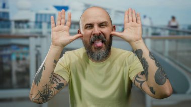 Sakallı ve dövmeli kel bir adam deniz kenarında bulanık bir geçmişi olan bir yolcu gemisinin güvertesinde komik suratlar yapıyor.