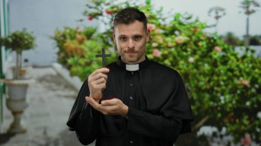 Bahçede haç tutan rahip düşünceli bir ifade ve papaz kıyafetiyle maneviyat ve sükunet duygusu sergiliyor.