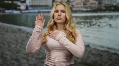 Kumsalda uzun saçlı, pembe gömlekli genç bir kadın yemin jesti yaparken deniz ve kumlu kıyı şeridi huzurlu bir zemin oluşturuyor..