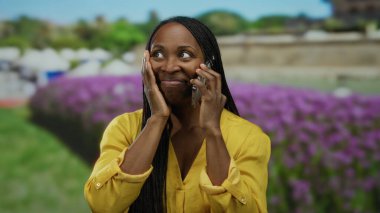 Parlak sarı gömlekli, neşeli bir şekilde konuşan bir kadın, güneşli bir parkta dikiliyor, arka planda mor çiçekler ve yeşillik var, mutluluk ve heyecan saçıyor..