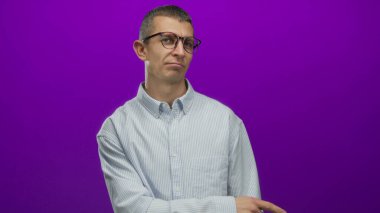 Gözlüklü beyaz bir adam, parlak mor bir arka plana karşı, çizgili bir gömlek giymiş, merak ya da endişeyle bakıyor..