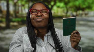 Baş ağrısı çeken kadın, Suudi pasaportunu açık hava parkı ortamında tutuyor. Stres ya da seyahat endişesini sakin doğal arka plan görünümünde dile getiriyor..