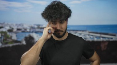 Sakallı genç adam limana yakın bir yerde duruyor. Arkaplanı açık gökyüzünün altında, gündüzleri siyah tişört giyerken.