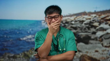 Gözlüklü ve steteskoplu genç adam kayalık bir plajda gülüyor mavi deniz arkaplanı ve berrak gökyüzü açık havada doğa ve sağlık hizmetlerini birleştiriyor..