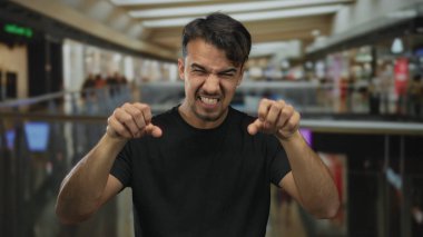 İspanyol asıllı genç bir adam kapalı bir alışveriş merkezinin arka planında neşeli ve anlamlı bir ortam yaratarak, kapalı bir alışveriş merkezinde jest ve şakacı bir gürleme yapıyor..