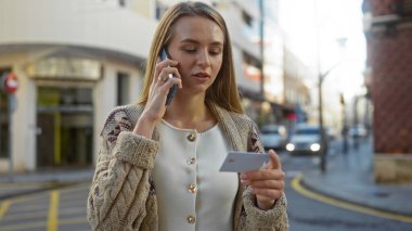 Kadın cep telefonuyla konuşuyor ve elinde kartla şehir caddesinde duruyor. Genç yetişkin kadın etkileşimine ve şehir atmosferine odaklanmış modern yaşam tarzını aktarıyor..