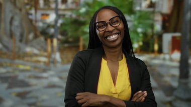 Şehir ortamında gülümseyen, gözlük takan, sarı gömlekli kıvırcık saçlı, kendine güveni ve mutluluğu şehrin arka planında gösteren bir kadın..