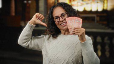 Şehirde orta yaşlı bir kadın Malezya ringgit notlarını kullanarak para gösteriyor, kendine güvenerek gülümsüyor ve finansal başarısını gösteriyor..