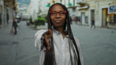 Şehirdeki sokakta el sıkışmak için elini uzatan kadın, şehir arka planında bulanık, açık havada dostça bir etkileşime işaret ediyor..