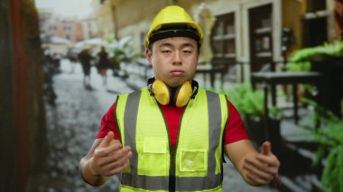 Şehir merkezinde arka planda bulanık duran, uykulu ve yorgun görünen, güvenlik yeleği ve sert şapka giyen bir adam çevredeki şehir hayatını gözler önüne seriyor..