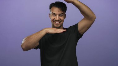 Genç İspanyol bir adam, neşeli ve yaratıcı bir stüdyo ortamında izole edilmiş mor bir arka plana karşı elleri ile bir çerçeve hareketi yaparken gülümsüyor..
