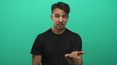 İzole edilmiş yeşil bir arka planda para için el kol hareketi yapan genç İspanyol bir adam, stüdyo ortamında ifadesini ve günlük kıyafetlerini vurguluyor.