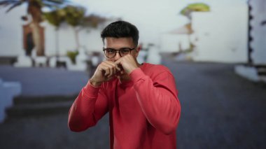 Kırmızı tişörtlü genç adam güneşli bir şehirde beyaz binalar ve palmiye ağaçlarıyla çevrili bir sokakta yumruklarıyla şakacı pozlar veriyor..